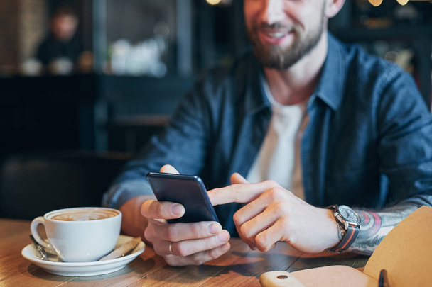 Руки людини в джинсовій сорочці слайд з пальцем на екрані його смартфон, біля чашки з кавою на дерев'яному столі
 - Фото, зображення