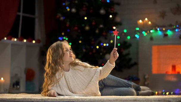 Cute girl holding magic wand, making wishes near Christmas tree, fairy-tale - Zdjęcie, obraz