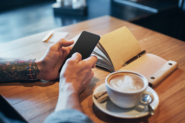 Uomo mani in denim scivolo camicia con dito sullo schermo il suo smartphone, vicino tazza con caffè sul tavolo di legno
 - Foto, immagini