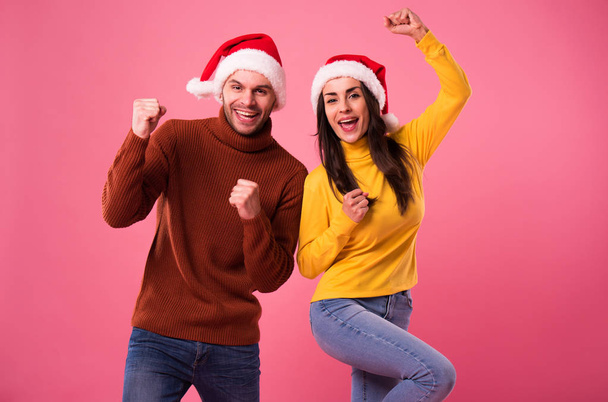 Excited and happy christmas couple! Young smiling beautiful couple in love in sweaters and santa hats have a fun and posing isolated on pink background - Foto, Bild
