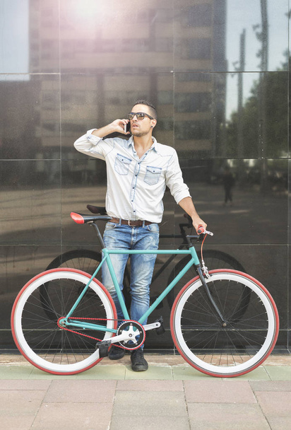 Young man talking on smart phone while standing by bicycle against black wall in the city - Fotografie, Obrázek