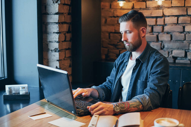 schöner junger Mann arbeitet am Laptop, während er Kaffee im Café genießt - Foto, Bild