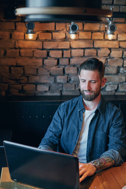 Joven guapo trabajando en el ordenador portátil mientras disfruta del café en la cafetería
 - Foto, Imagen