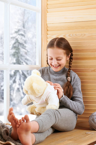 Gyermek lány ül a ablakpárkányon, és a játék-játék medve. Gyönyörű kilátással az ablakon - süt a nap, a hó és a tél. - Fotó, kép
