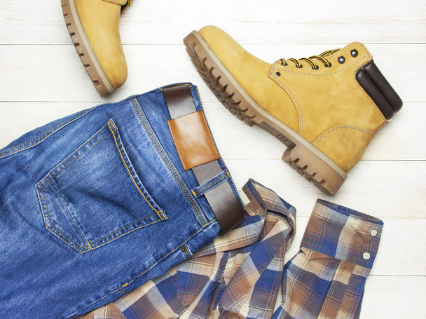 Мужская повседневная одежда, жёлтые рабочие сапоги из натуральной кожи нубака, синие джинсы, клетчатая рубашка и коричневый пояс на деревянном белом фоне. Традиционная повседневная обувь, молодежный стиль
 - Фото, изображение