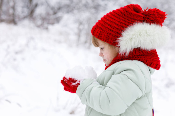 A little cute girl in bright clothes with funny emotions on her face is standing sidelong and holding a heap of snow in her hand at winter park background. - Photo, image