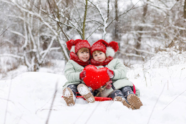 Gêmeos menina estão sentados no chão nevado e segurando juntos um coração vermelho brilhante em suas mãos.O conceito é Dia de São Valentim
 - Foto, Imagem
