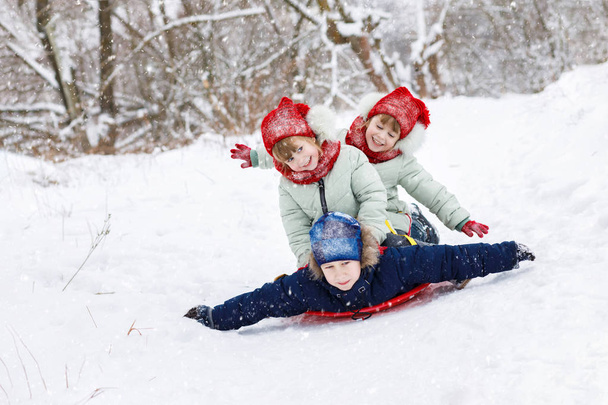 Ευτυχώς γέλιο δίδυμα κορίτσια κάθονται στην πλάτη του αγοριού τοποθέτηση πλαστικής πλάκας και έλκηθρο από τον λόφο μεγάλο χιόνι. Η έννοια είναι η υπαίθρια υπαίθρια δραστηριότητα χειμώνα. - Φωτογραφία, εικόνα