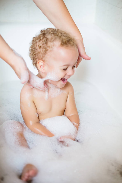 Closeup two year old child having fun with foam by bathing in bathtub. The symbol of purity and hygiene education. - Photo, Image