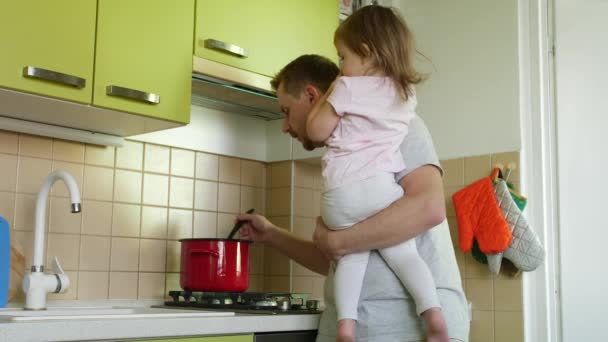 Father's day. Cooking father with small daughter in hands yelling at son for homework in kitchen. Dad making a meal, taking care of small girl and berates for lessons elder boy. 2 children claim parent's attention at home. Paternity leave with kids - Footage, Video