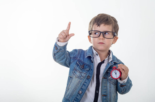 Happy young boy in white t-shirt and jeans jacket thinking and looking up with red clock pointing finger up on white background. Kid with alarm clock - Foto, imagen