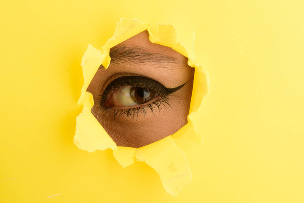Eye close up. Spying, secret. Beautiful female eye with simple make up is looking at camera through the hole in yellow paper. Female eye seen through the torn paper. Curiosity from young girl - Photo, Image