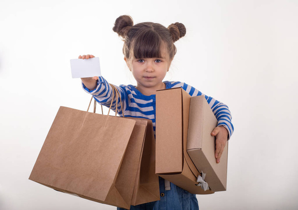 Симпатична щаслива дитина тримає дисконтну білу картку, сумки і коробки в руках. Малюк з кредитною карткою. Маленька дівчинка показує порожній купон, копіює простір. Ізольовані
 - Фото, зображення