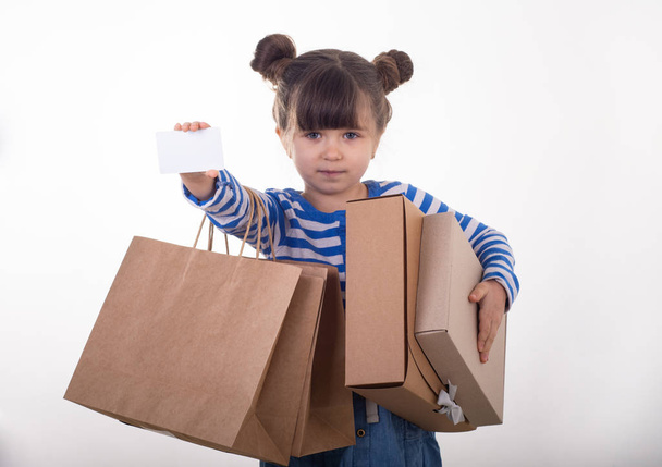 Bonito criança feliz segurando desconto cartão branco e sacos de compras e caixas em suas mãos. Um miúdo com cartão de crédito. Menina mostrando cupom vazio, espaço de cópia. Isolados
 - Foto, Imagem