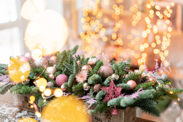 Beautiful festive arrangement of fresh spruce, pink ornamentals in a rustic wooden box box. Christmas mood. Garland bokeh on background. - Фото, зображення