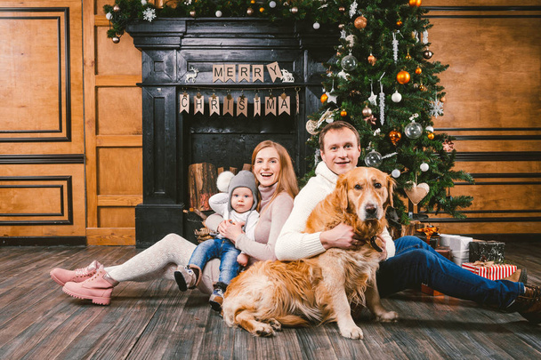 Tema Noel ve yeni yıl aile çevresi. Genç beyaz aile ile 1 yaşındaki çocuk köpek doğurmak Labrador Golden Retriever ahşap zemin oturma odasında şömine Noel ağacı yakınındaki ev üzerinde oturan. - Fotoğraf, Görsel
