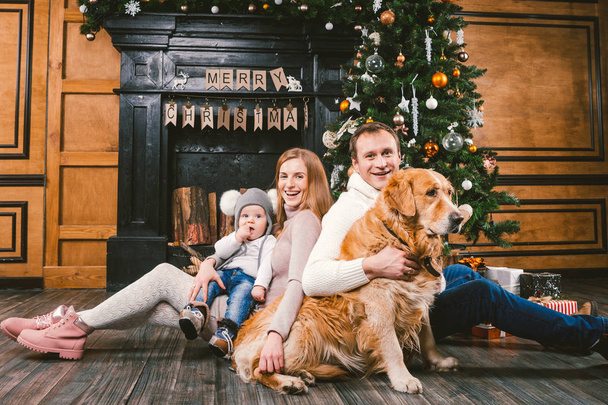 Téma karácsonyi és újévi családi körben. Fiatal kaukázusi család 1 éves gyermek kutya tenyészt Labrador Golden Retriever ül otthon, a nappaliban kandalló karácsonyfa közelében fa padló. - Fotó, kép