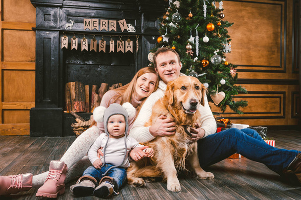 Thema Weihnachten und Neujahr Familienkreis. junge kaukasische Familie mit 1-jährigem Kind Hunderasse Labrador Golden Retriever sitzt auf Holzboden zu Hause im Wohnzimmer in der Nähe Kamin Weihnachtsbaum. - Foto, Bild