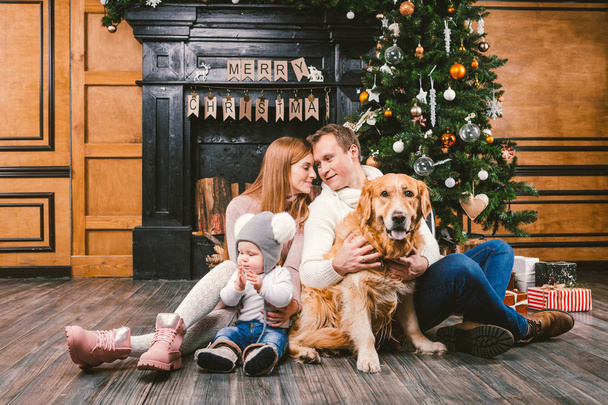 テーマ クリスマスと新しい年の家族の輪。若い白人家族は 1 歳の子の犬と繁殖する木製の床のリビング ルームの暖炉クリスマス ツリーの近くで家の上に座ってラブラドール ゴールデンレトリバー. - 写真・画像