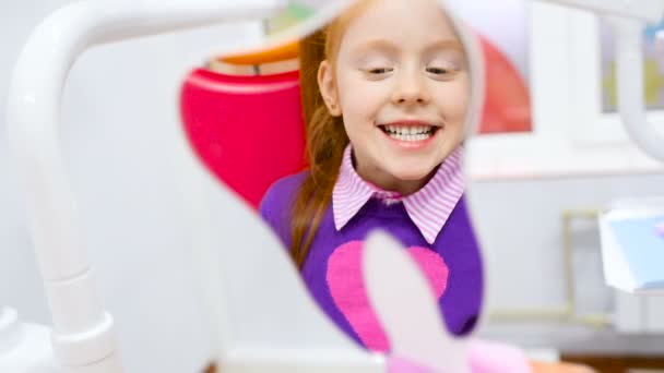 a little cute girl with red hair admires her healthy teeth in the mirror sitting in the dental chair. - Footage, Video