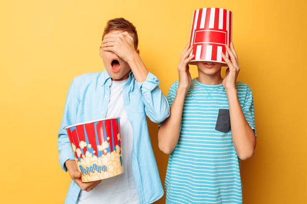 Image of two frightened teenagers, guys watching a horror movie and hiding behind a bucket of popcorn - Фото, изображение