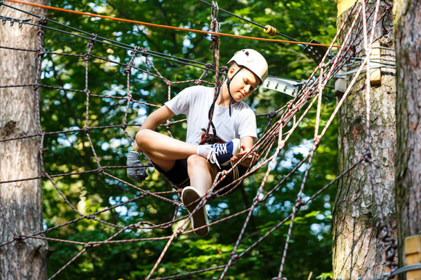 Child in forest adventure park. Kid in orange helmet  and blue t shirt climbs on high rope trail. Agility skills and climbing outdoor amusement center for children. young boy plays outdoors - Foto, Bild