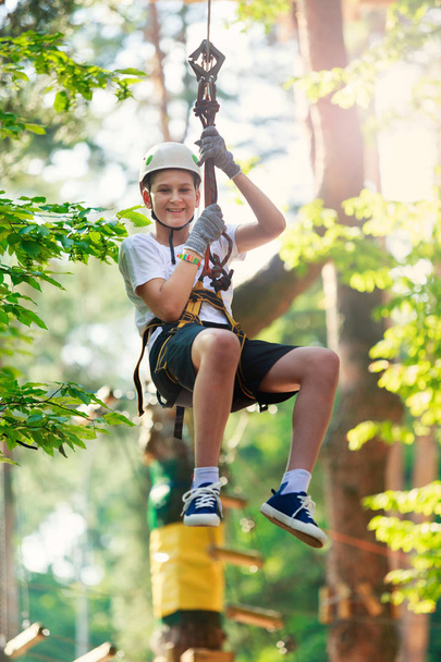 Child in forest adventure park. Kid in white helmet and white t shirt climbs on high rope trail. Agility skills and climbing outdoor amusement center for children. young boy plays outdoors - Photo, Image