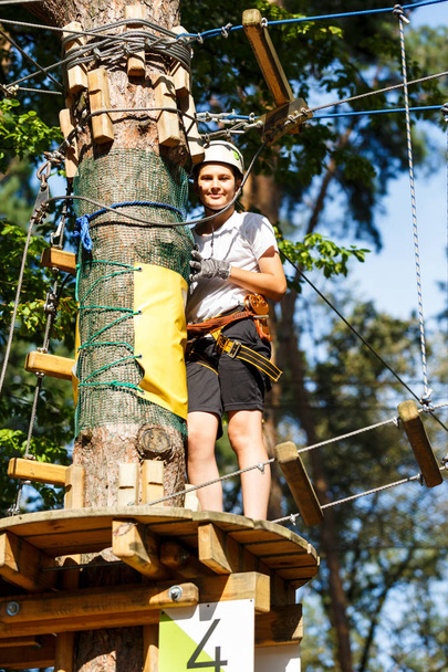 Child in forest adventure park. Kid in white helmet and white t shirt climbs on high rope trail. Agility skills and climbing outdoor amusement center for children. young boy plays outdoors - Φωτογραφία, εικόνα