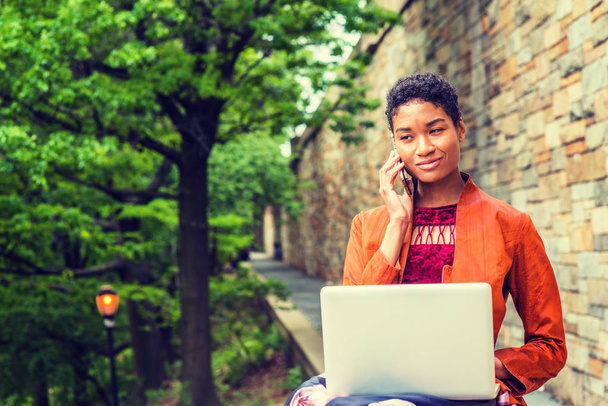 Young African American Woman studying, working on laptop computer, talking on cell phone outside in New York City, wearing orange red jacket, flower patterned skit, sitting by trees, walls on campus - Fotó, kép