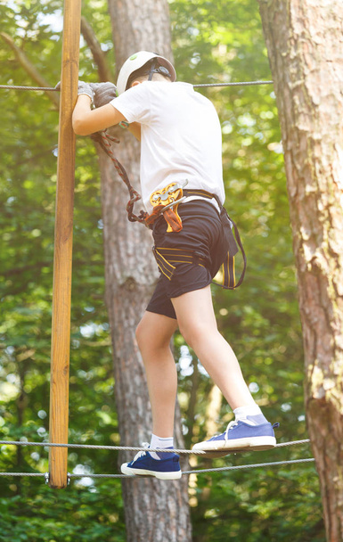Child in forest adventure park. Kid in white helmet and white t shirt climbs on high rope trail. Agility skills and climbing outdoor amusement center for children. young boy plays outdoors - Photo, Image