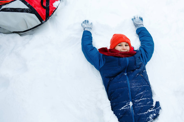 schattige jonge jongen in hoed blauw jasje houdt en speelt met sneeuw, heeft fun, glimlacht, maakt sneeuwpop in winter park. Actieve levensstijl, winter activiteit, buiten winter spelletjes, sneeuwballen.  - Foto, afbeelding