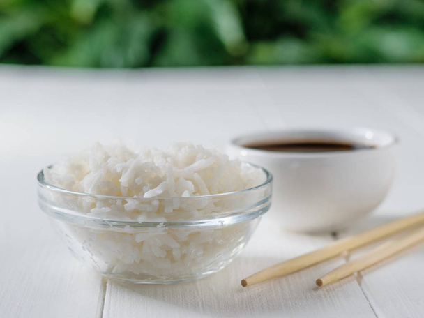 Glass bowl with white long grain rice, bowl with soy sauce and wooden sticks on white table on green tree background. Natural diet food. - Photo, Image