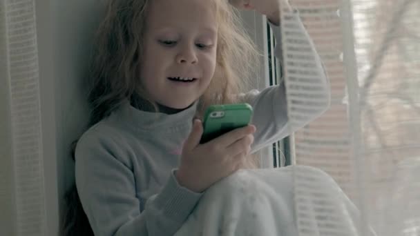 happy little girl with wavy red hair sitting on the windowsill, covering a blanket and using the phone, talking, video calling, close-up portrait - Materiał filmowy, wideo