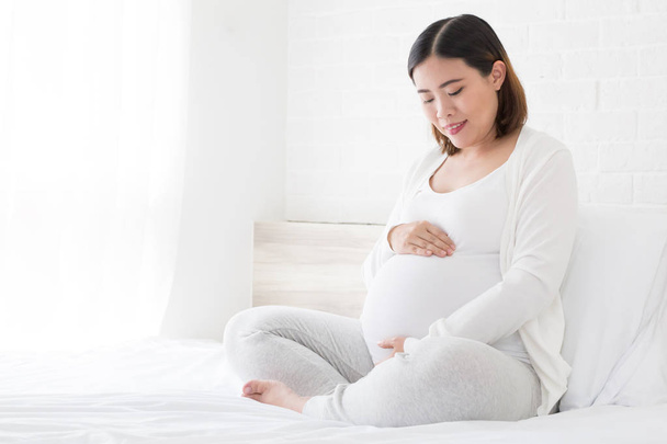 Happy asian pregnant woman sitting on bed and touching her belly at home - Foto, Imagem