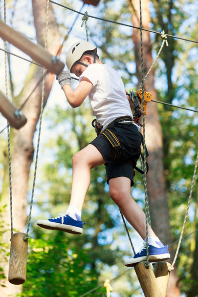 Child in forest adventure park. Kid in white helmet and white t shirt climbs on high rope trail. Agility skills and climbing outdoor amusement center for children. young boy plays outdoors - Fotoğraf, Görsel