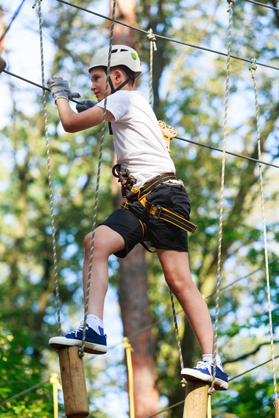 Child in forest adventure park. Kid in orange helmet  and blue t shirt climbs on high rope trail. Agility skills and climbing outdoor amusement center for children. young boy plays outdoors - Photo, Image