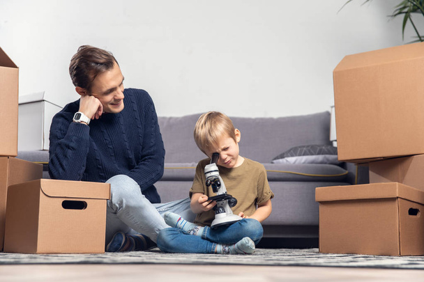 Photo of happy father and small son with microscope sitting on floor among cardboard boxes - 写真・画像