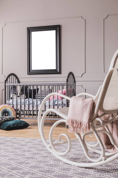 White rocking chair with pastel pink blanket in elegant little baby bedroom with grey wooden crib and mockup poster on the wall - Photo, image
