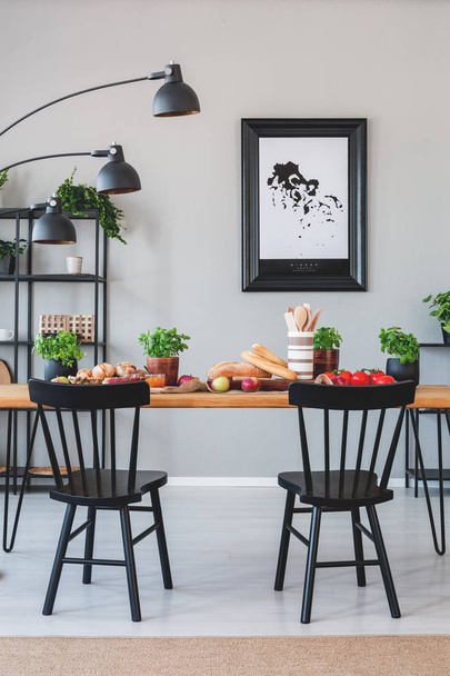 Lamp above black chairs and wooden table with food in grey dining room interior with poster. Real photo - Фото, изображение