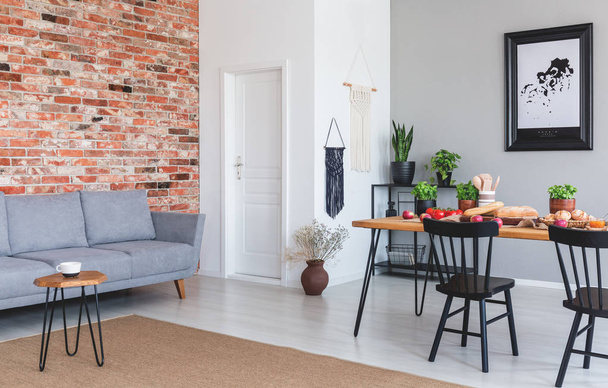 Grey sofa against red brick wall in flat interior with poster and black chairs at dining table. Real photo - Foto, Imagem