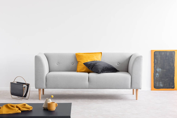 Yellow and black pillows on grey settee in spacious living room interior with blackboard in orange frame and magazine rack on the floor - 写真・画像