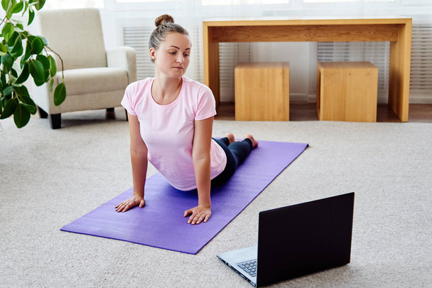 junge Frau praktiziert Yoga mit Laptop zu Hause drinnen, Kopierraum. Mädchen in Kobra-Pose, volle Länge. Entspannung und Yoga. Wellness und gesunder Lebensstil. bhujangasana - Foto, Bild