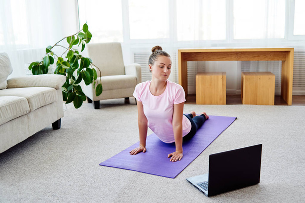 Young woman practicing yoga with laptop at home indoor, copy space. Girl practicing cobra pose, full length. Relaxing and doing yoga. Wellness and healthy lifestyle. Bhujangasana - Foto, Bild