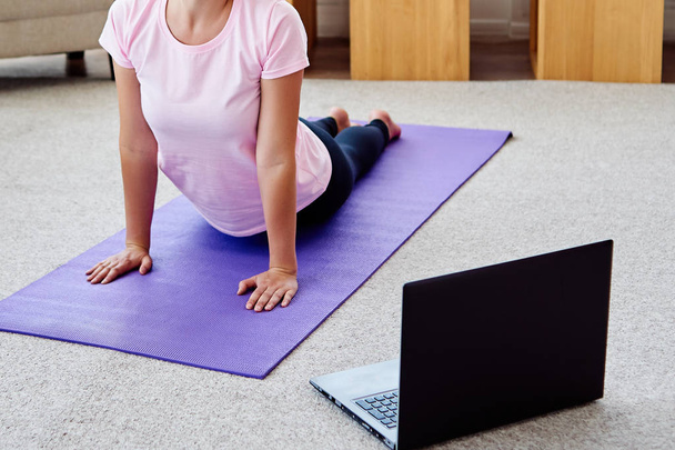 Young woman practicing yoga with laptop at home indoor, copy space. Girl practicing cobra pose. Relaxing and doing yoga. Wellness and healthy lifestyle. Bhujangasana - Zdjęcie, obraz
