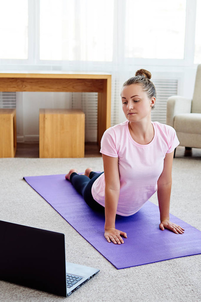 junge Frau praktiziert Yoga mit Laptop zu Hause drinnen, Kopierraum. Mädchen in Kobra-Pose, volle Länge. Entspannung und Yoga. Wellness und gesunder Lebensstil. bhujangasana - Foto, Bild