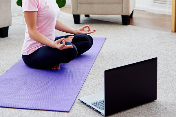 Girl sitting in lotus position and meditating at home interior, copy space. Padmasana. Woman practicing home yoga with laptop. Hands in meditating gesture. Freedom concept - Photo, image