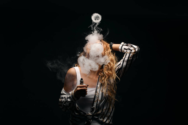 Redhead woman vaping electronic cigarette with smoke on black background closeup. Young woman smoking e-cigarette to quit tobacco. Vapor and alternative nicotine free smoking concept, copy space  - Zdjęcie, obraz