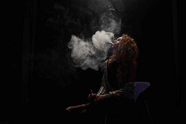Redhead woman vaping electronic cigarette with smoke on black background closeup. Young woman smoking e-cigarette to quit tobacco. Vapor and alternative nicotine free smoking concept, copy space  - Фото, изображение