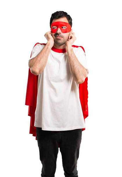 Superhero man with mask and red cape covering both ears with hands. Frustrated expression on isolated white background - Foto, Bild