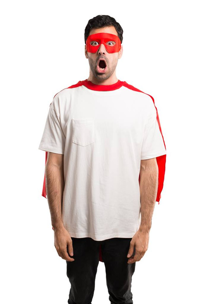 Superheldenmann mit Maske und rotem Umhang mit überraschendem und schockiertem Gesichtsausdruck. klaffend, weil sie gerade mit einem Geschenk auf isoliertem weißem Hintergrund überrascht haben - Foto, Bild